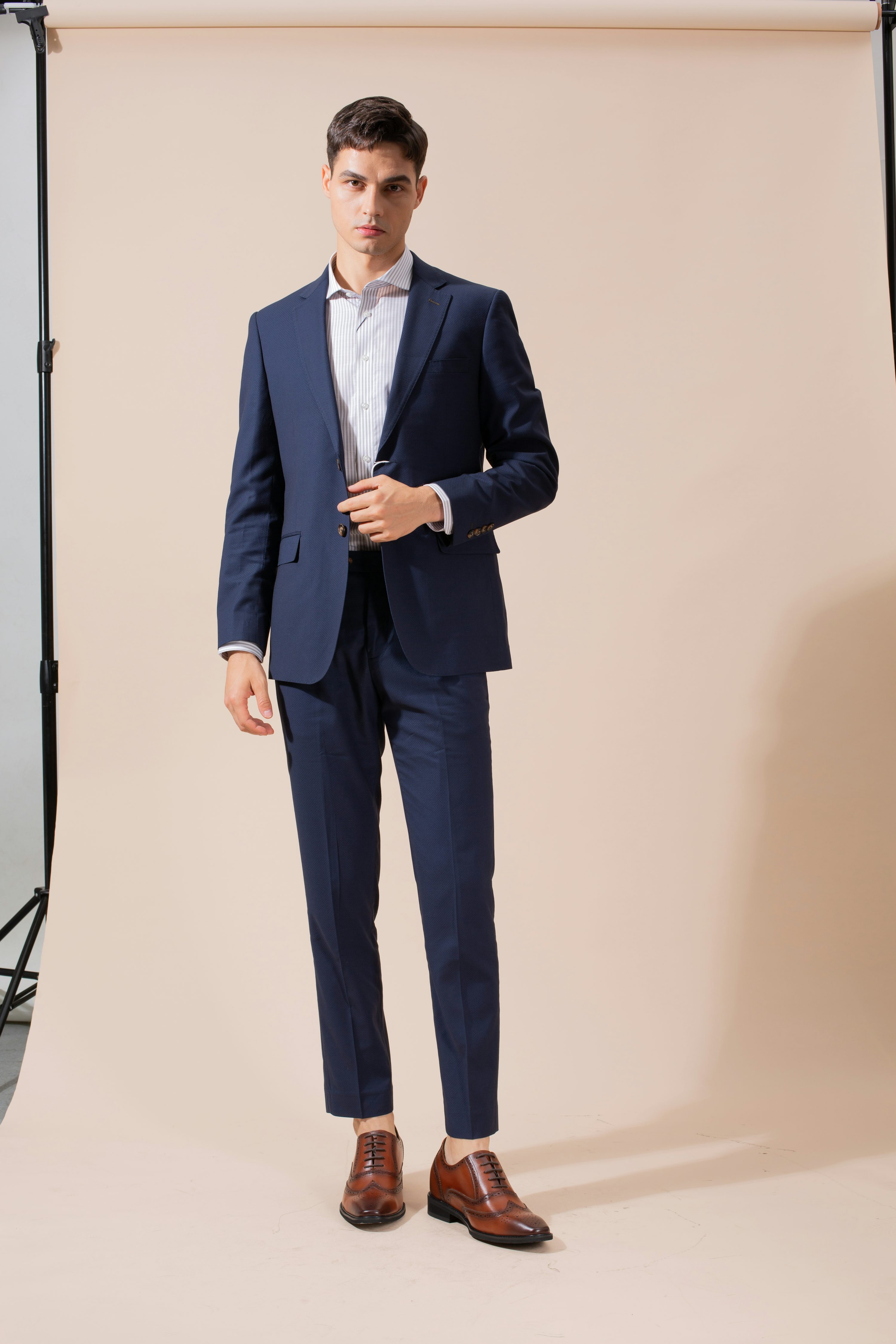 a fullbody shot of a handsome, confident man in a navy blue suit and white shirt. he is standing against a light beige background, looking directly at the camera. his shoes are brown oxfords, matching his sophisticated and elegant look.  the setting appears to be a professional studio.