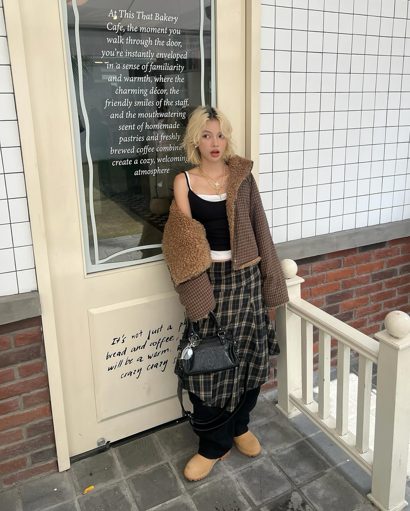 a young woman stands in front of a cafe, which is described on a sign as having a cozy and welcoming atmosphere. she is wearing a brown teddy coat over a black tank top and a plaid skirt. she also has a black handbag and brown boots. her hair is blonde. the overall style is autumnal and chic.