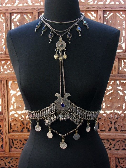 the image shows a silver necklace and body jewelry displayed on a black mannequin. the necklace consists of multiple strands and pendants, some with coins. the body jewelry wraps around the torso, also with coins and decorative elements. the background features a wooden screen with intricate patterns.