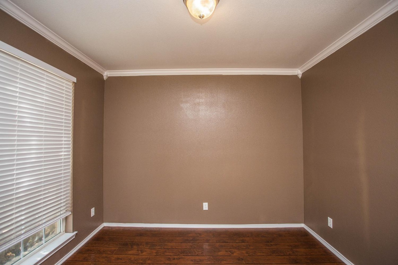 a medium shot of an empty room with brown walls and dark wood floors. there is a window with closed blinds to the left, and a light fixture on the ceiling. the room has white crown molding and baseboards, and there are electrical outlets on the walls. the wall behind the view is empty.