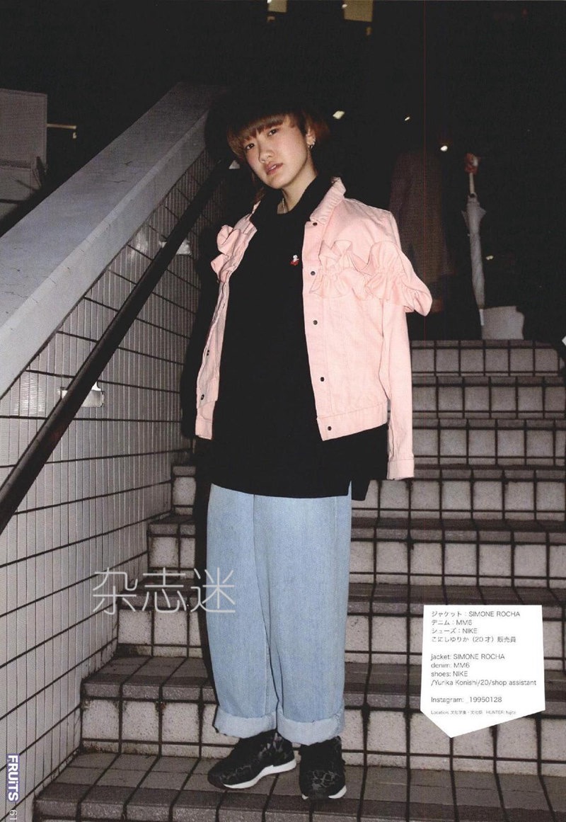 a young man is posing on a city staircase at night. he is wearing a pink oversized denim jacket with ruffled details on the shoulders, a black sweatshirt, baggy lightwash jeans, and black sneakers with white accents.  the photo has a grainy texture and a vintage aesthetic, reminiscent of 90s japanese street style. japanese characters are visible in the image.