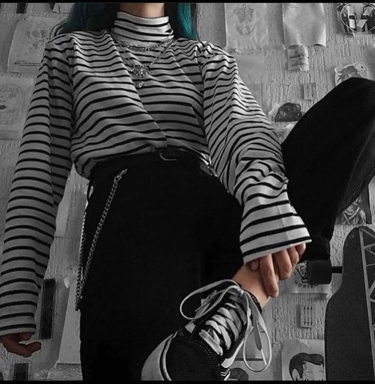 a young woman is wearing a black and white striped longsleeve shirt, black pants, and black sneakers. she is sitting with her legs crossed and holding a skateboard. the photo is taken in a black and white aesthetic. the woman looks cool and edgy.