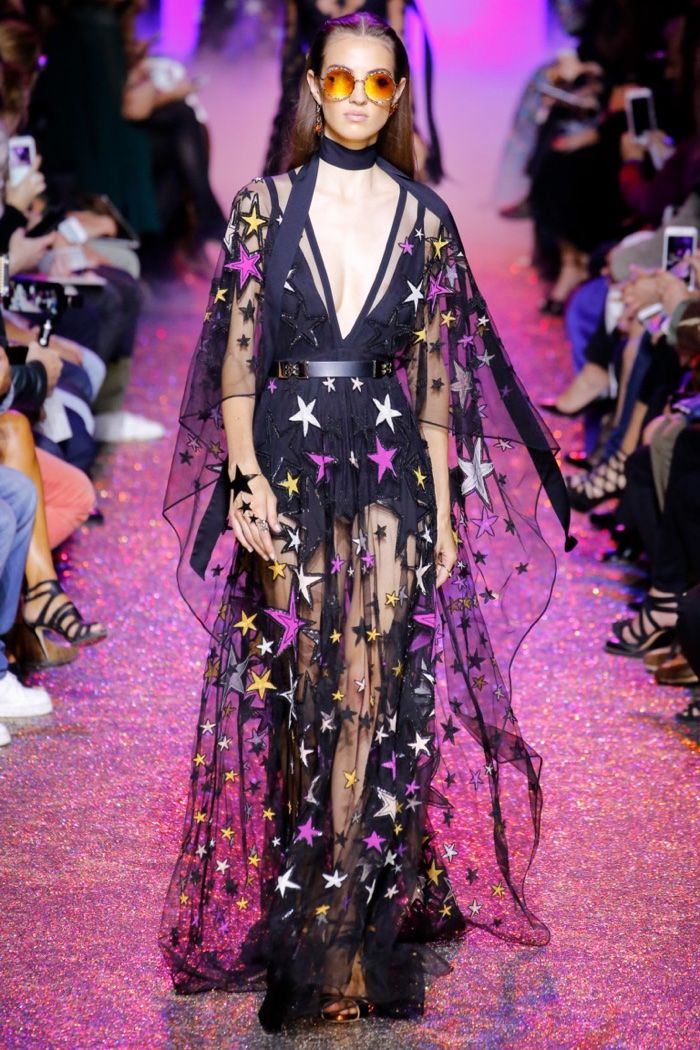 a model walks the runway in a black sheer gown adorned with stars of various colors and sizes. the dress is layered over a black bodysuit, and the model is accessorized with sunglasses, a black belt, and delicate jewelry. the background is filled with glitter and people watching the show. the model's elegant pose and the overall look exude high fashion and glamour.