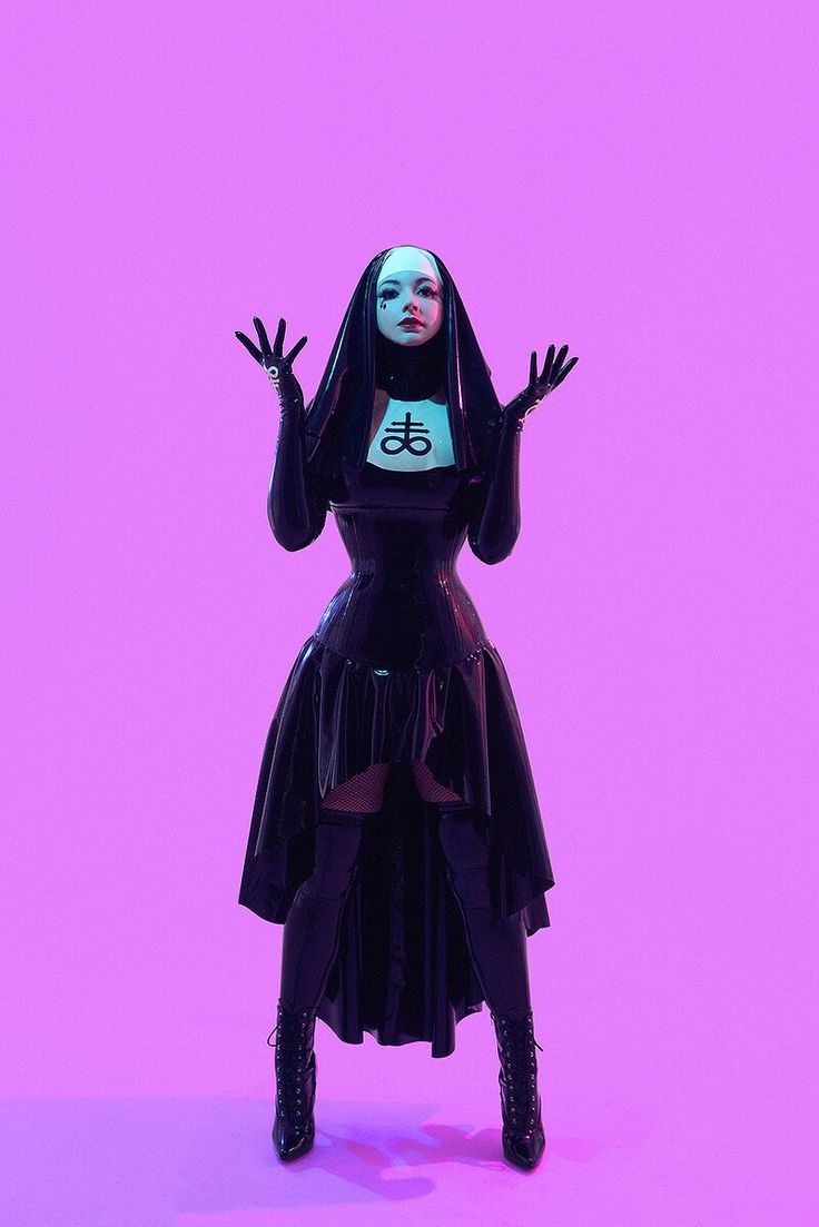 a woman dressed in a gothic style, posing in a black latex dress and boots, with a pentagram on her collar. she is standing in a studio with a pink background and looking at the camera.