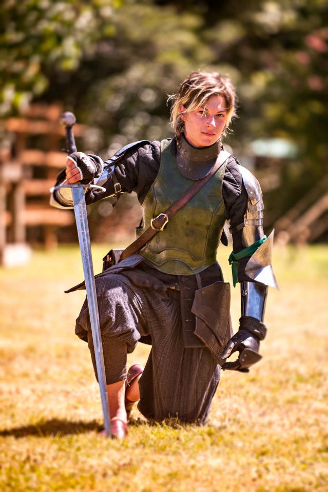 a female knight dressed in full armor, kneels in a grassy field, holding a sword in her right hand.  she is looking directly at the camera with a determined expression on her face.  she is a strong, powerful warrior ready for battle.