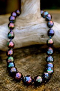 a black pearl necklace with an iridescent finish is displayed on a piece of driftwood. the pearls are a deep, rich black with hints of green, blue, and purple shimmering in the light. the driftwood provides a natural and rustic backdrop for the elegant necklace.