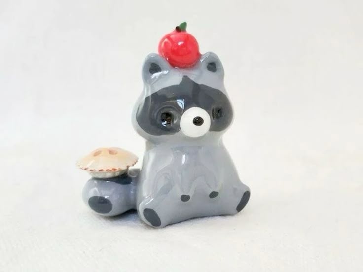 a cute ceramic figurine of a gray raccoon is sitting with a red apple on its head and a mushroom on its tail. the raccoon has a black mask and a single, large white eye. the figurine is whimsical and detailed, suggesting it is a collectible art piece.