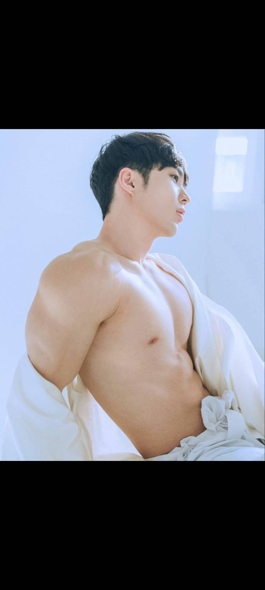 the image shows a shirtless, muscular asian man with short dark hair. he is posing in profile, looking to his right, with his body slightly angled. he is partially draped in white cloth that is loosely wrapped around his torso and lower body.  the lighting is bright and soft, creating a light and airy feel. the overall aesthetic is sensual and suggestive, highlighting the man's physique.