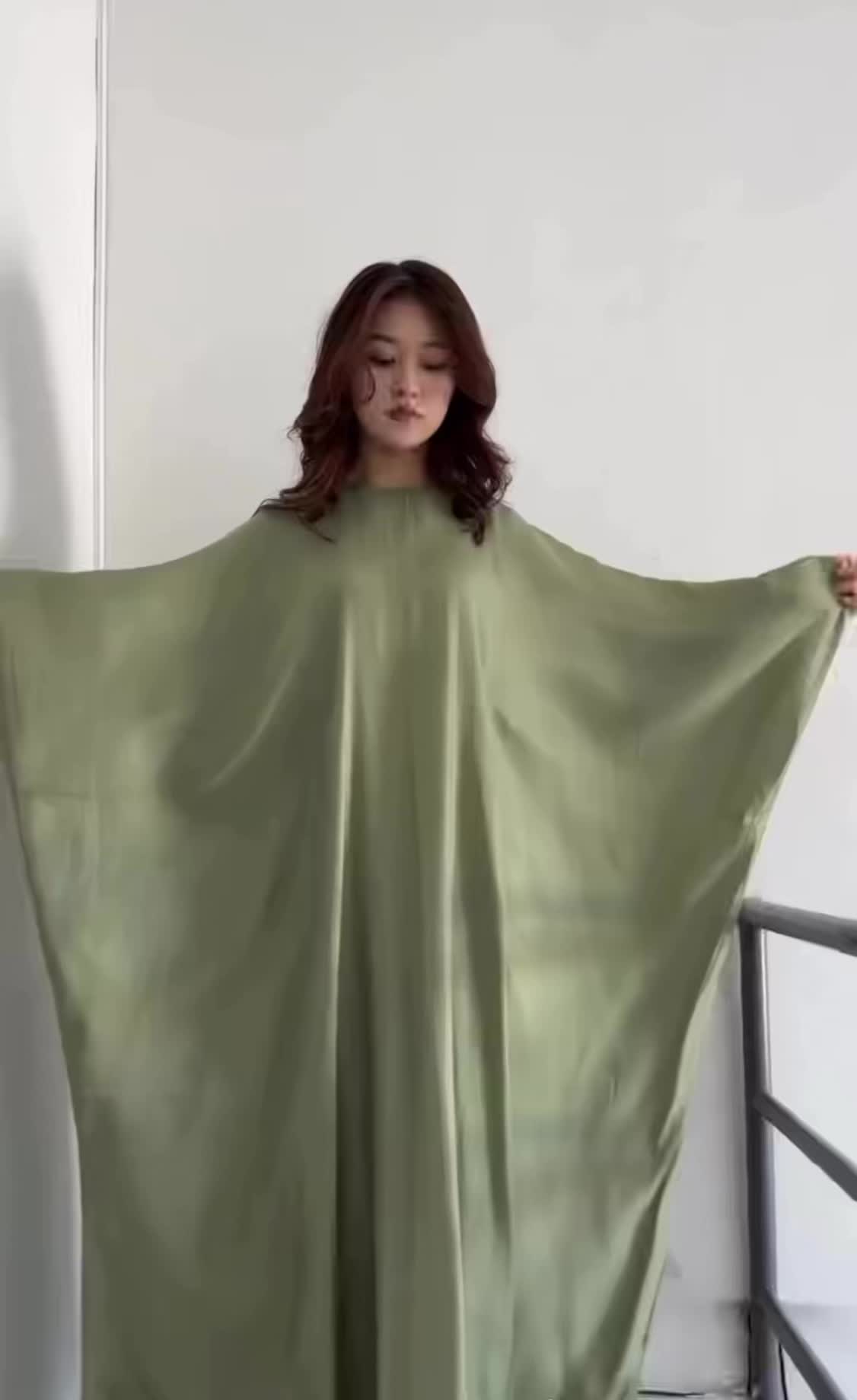a woman wearing a light green dress stands in front of a white wall. she is wearing an oversized dress, as she has her arms outstretched. the dress has a slightly shiny appearance and the woman appears to be in her twenties. a black railing is visible next to her.