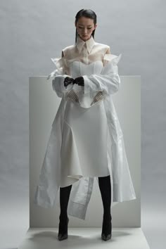 a woman is standing in a studio wearing a white dress with a large white skirt, a white shirt, and black shoes. she has her hair pulled back. she is looking down at her hands.