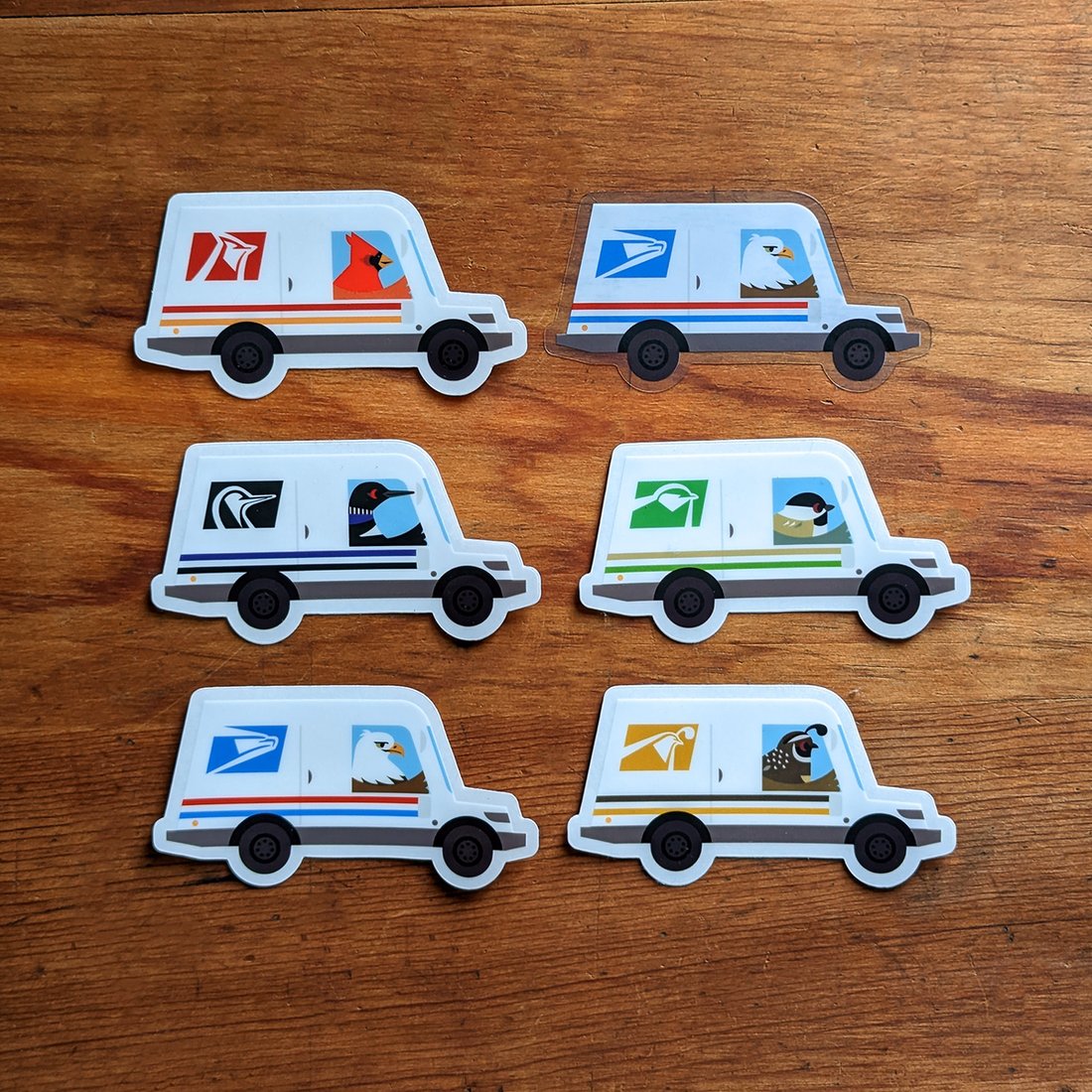 the image shows six different stickers of stylized usps mail trucks. each truck features a different bird species sitting in the passenger seat: a cardinal, an eagle, a loon, a puffin, a quail, and another bird. the stickers are arranged in two rows of three on a wooden surface. the style of the stickers is clean and simple, using bright colors and flat illustrations.  the overall aesthetic suggests a playful and somewhat whimsical representation of mail delivery.