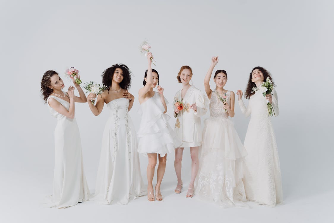 a group of six diverse bridesmaids in elegant white dresses stand together against a white background, each holding a bouquet of flowers.  they are smiling and appear happy, some are playfully posing with their bouquets.  the overall feel of the image is joyful and celebratory.
