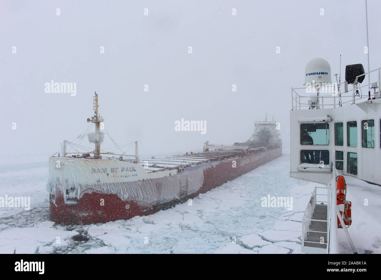 a view from the bridge of a ship as it navigates through icy waters in the great lakes.  a large cargo ship, covered in ice, is in the distance, while the viewer's ship is in the foreground.