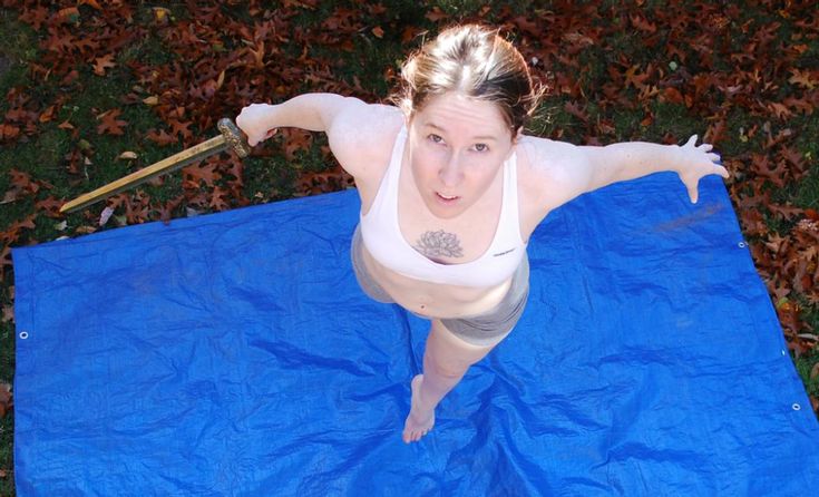 a young woman in a white sports bra and gray shorts is lying on her stomach on a blue tarp, looking down at a sword she is holding in her hand. the ground is covered in brown leaves and green grass. it is an outdoor setting, and it seems like the woman is dressed as a warrior.