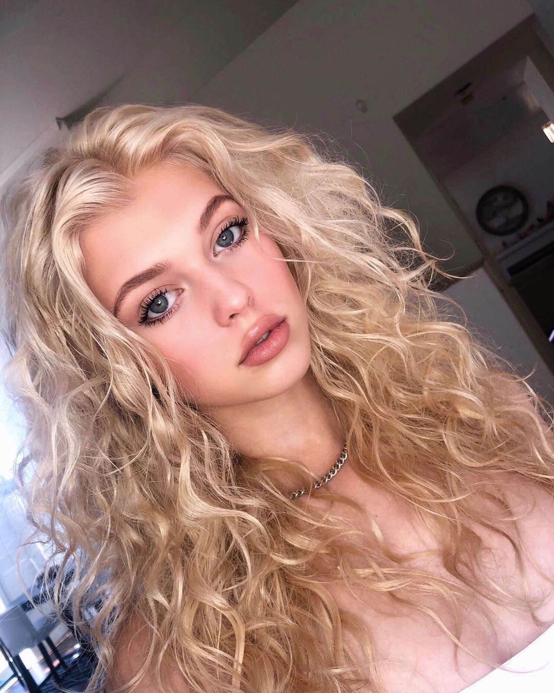 a closeup portrait of a young woman with long, curly blonde hair, blue eyes, and nude lipstick. she is wearing a silver necklace and a white top. the background is slightly blurred, showing a glimpse of a room.