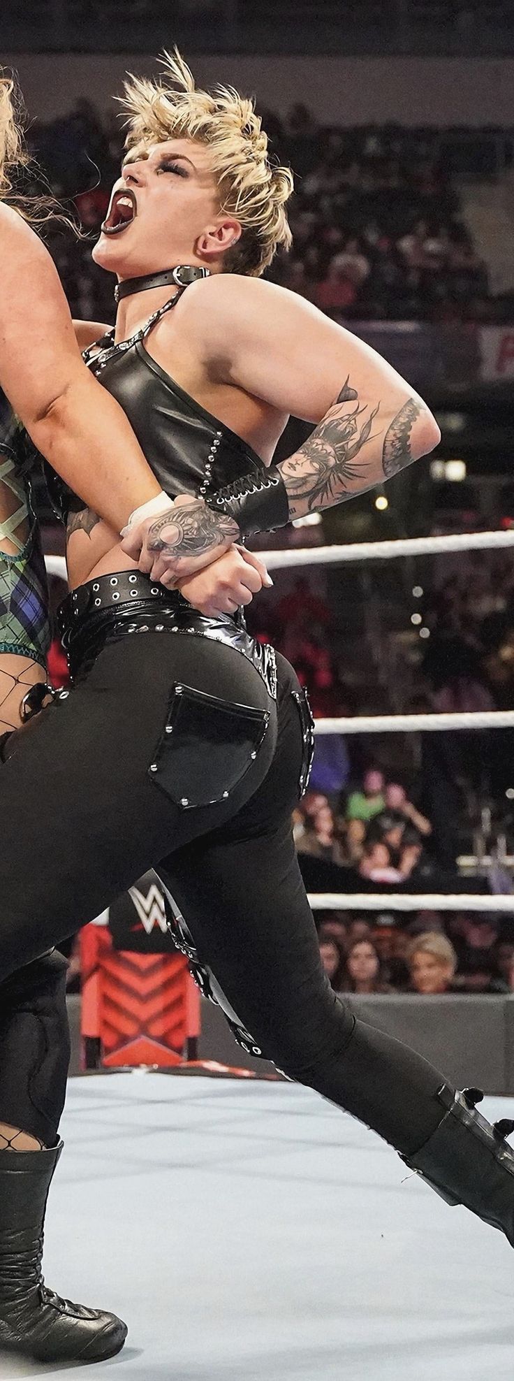 a female wrestler with blonde hair is standing in a wrestling ring. she's wearing black leather pants and boots and has a tattoo on her arm. the image is taken from a low angle.  she appears to be in midpose or midmove. the background is blurred, likely due to the action of the match.
