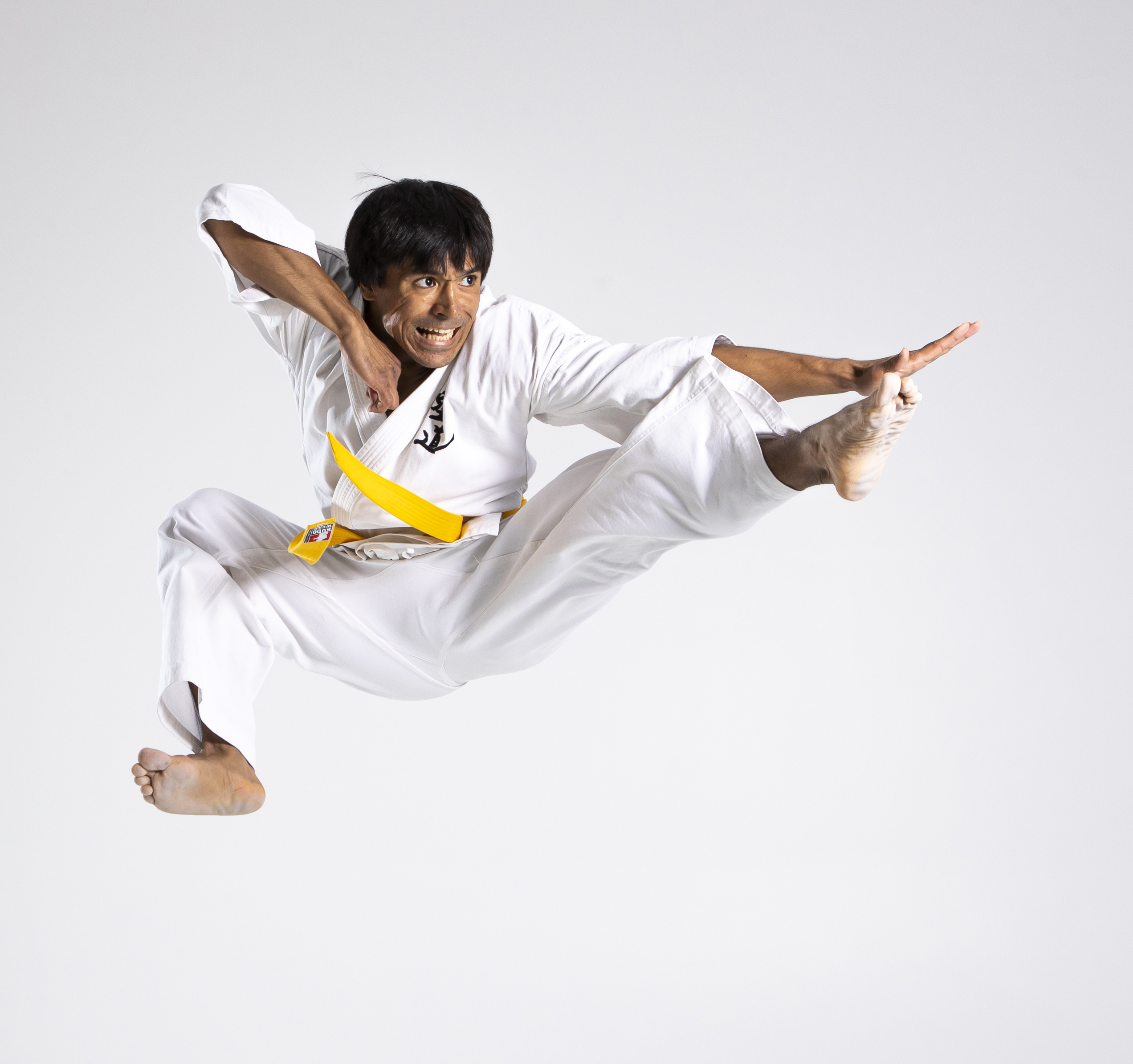a man wearing a white karate uniform with a yellow belt is performing a flying kick against a white background. he is midair, with one leg extended and the other bent, his arms are outstretched for balance, and he has a determined expression on his face.
