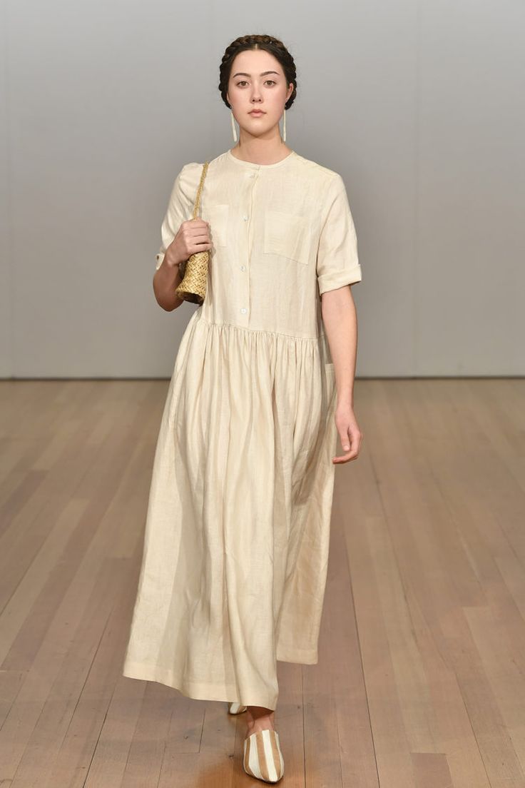 the image shows a fashion model walking down a wooden runway. she is wearing a light beige linen maxi dress with short sleeves, a buttondown front, and a gathered waistline. the dress has a simple, elegant style. she carries a small, lightcolored woven straw bag and wears beige striped mules. her hair is styled in neat braids, completing the minimalist and chic look. the background is a plain, lightcolored wall and wooden floor, focusing attention on the model and her outfit.  the overall aesthetic is casual yet refined, suggesting a summer or spring collection.