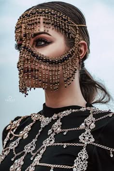 a glamorous portrait of a woman with long dark hair and striking makeup. she wears a intricate gold beaded headpiece that covers her face, resembling a mask, leaving only her eyes visible. the headpiece is adorned with delicate chains and embellishments, creating a sense of both mystery and elegance.  she also wears a  layered necklace made of silver chains and intricate designs, which  extends down her shoulders. the image has a dramatic and alluring aesthetic, highlighting the woman's beauty and the exquisite detail of her jewelry. the blurred background draws attention to the subject and enhances the overall mood of the photograph.