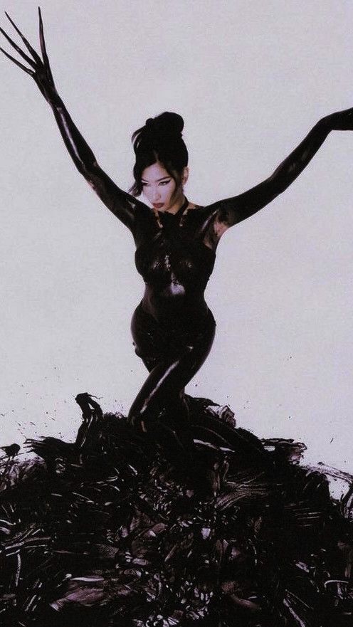 this is a dramatic portrait of a woman in a black bodysuit, posed with one arm and leg extended, and the other hand posed with fingers extended as if reaching out to the sky. her body is painted black, as is the background and the floor, which resembles a messy pool of paint. she looks mysterious and alluring, a dark and edgy beauty.