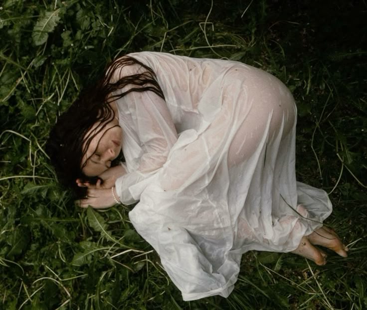 a woman wearing a wet, white dress is curled up on the ground in a patch of grass. she is lying down with her eyes closed and her dark hair spread around her head. her bare feet are visible. the scene evokes a sense of melancholy and vulnerability.
