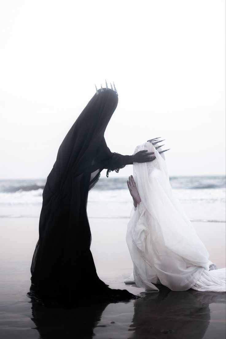 two women, one in a black gown and crown and the other in a white gown, are standing on a beach, looking at the ocean. the woman in the black gown has her back turned to the camera, while the woman in the white gown is looking at the ocean. both women are wearing  crowns. the background is an ocean and beach. the overall tone of the image is dark and mysterious, but also beautiful. the two figures contrast against the soft background, creating a dramatic scene that appears gothic and stylish.