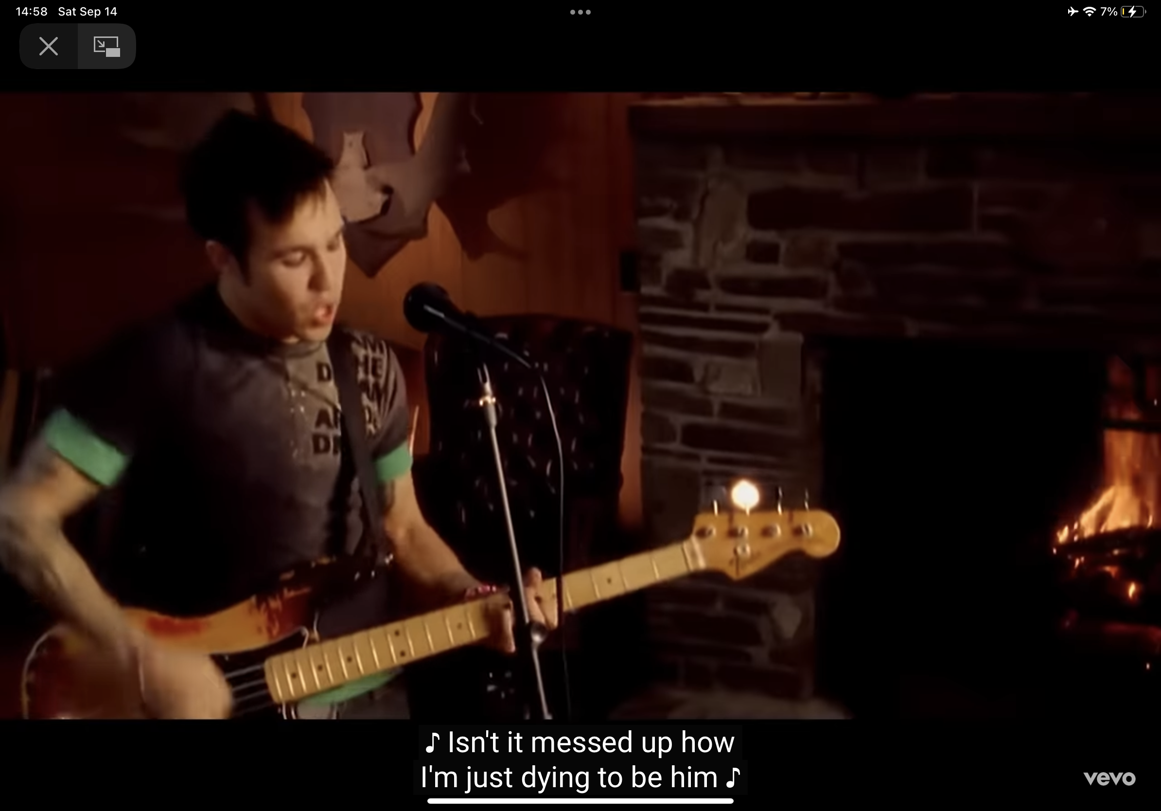 a man with short dark hair and tattoos is playing a bass guitar and singing. he is wearing a green tshirt and standing in a dimly lit room with a stone fireplace in the background. the image has a warm tone to it.