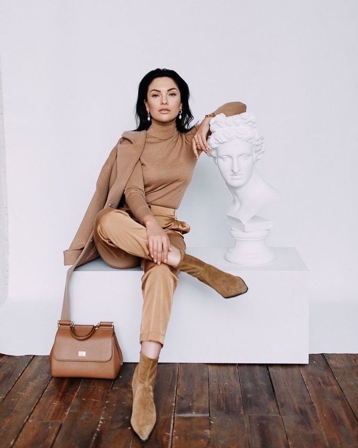 a fashionable brunette woman is sitting elegantly on a white cube in front of a white bust sculpture. she is wearing a camelcolored turtleneck sweater, satin trousers, and ankle boots.  she is holding a dolce & gabbana handbag. the background is a minimalist white studio setting with wooden floors. the overall style is sophisticated and chic.