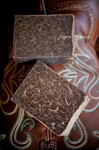 a closeup shot of two bars of brown soap, likely handmade, resting on a piece of leather with a western design. the soap has a rustic and natural appearance, suggesting it's crafted with organic ingredients. the overall composition is warm and inviting, highlighting the beauty and craftsmanship of handcrafted products.