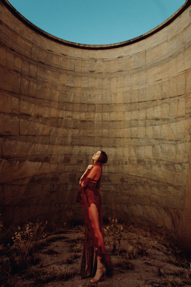 a woman in a long, flowing red dress is standing in the center of a circular stone structure. the structure has a large opening at the top, allowing natural light to illuminate the scene. the woman is posing with her eyes closed and her arms crossed in front of her chest. the overall tone of the image is soft and ethereal.