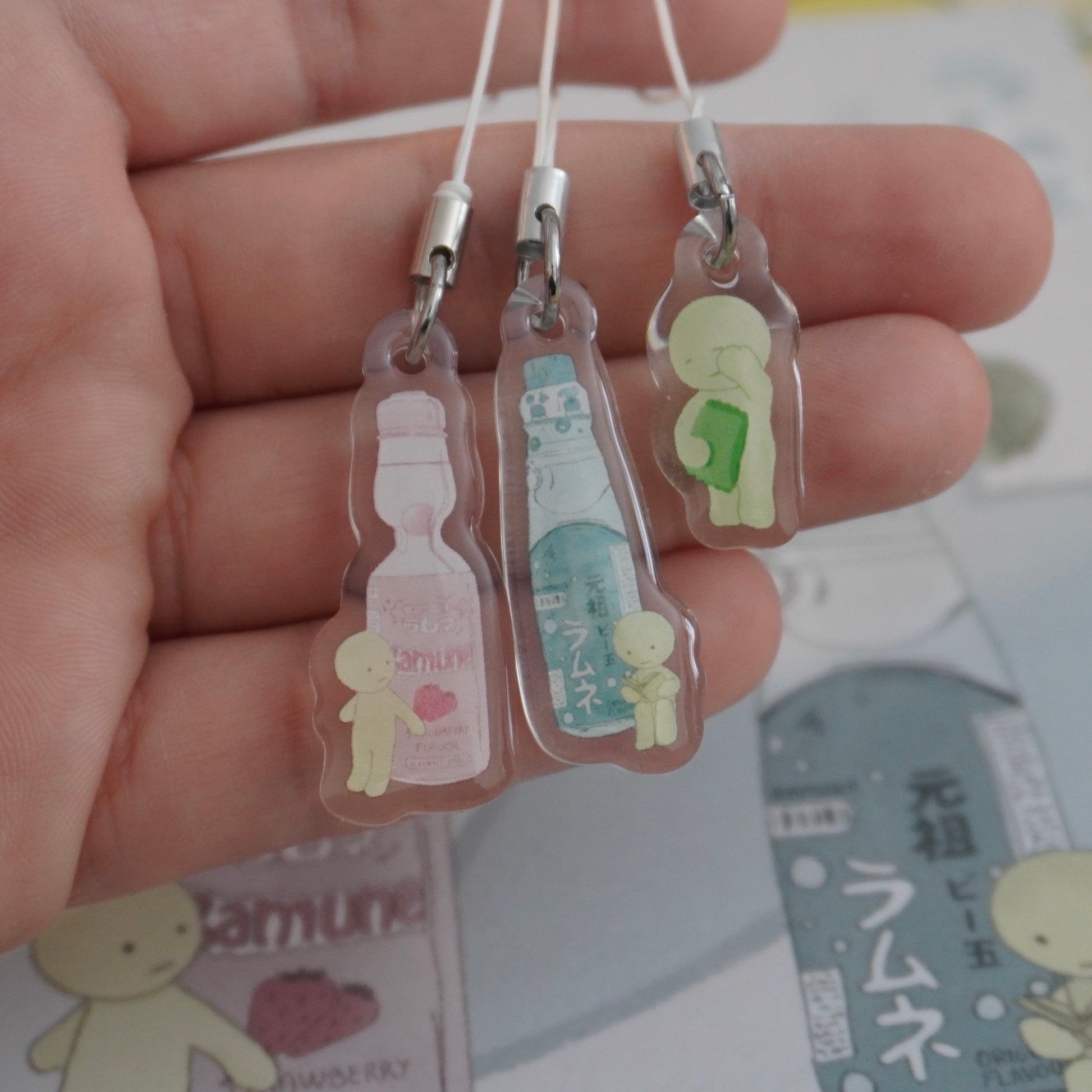 a closeup shot of a hand holding three handmade resin keychains. each keychain features a small, cute character and a ramune bottle, a type of japanese soda. the bottles are depicted in pastel colors, with pink, green and blue versions. the keychains are transparent, allowing the images inside to be clearly seen. the hand is gently holding the charms, showcasing their delicate nature.  the background includes part of a blurry image that appears to be related to ramune products.