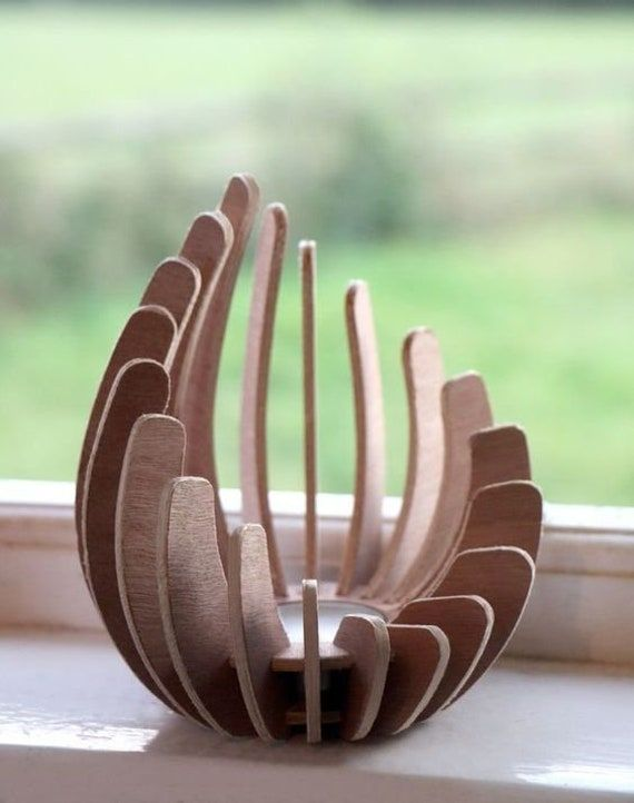 a light brown wooden candle holder sits on a white windowsill in front of a window. the candle holder is ovalshaped and made of thin strips of wood that are arranged in a spiral pattern. the background is a blurry green field.