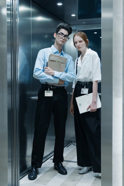 a full shot of a man and a woman standing in an elevator. they are both dressed in business attire. the man is holding a folder, while the woman is holding a laptop. they appear to be colleagues or coworkers. the elevator is modern and stylish, with a metallic interior.