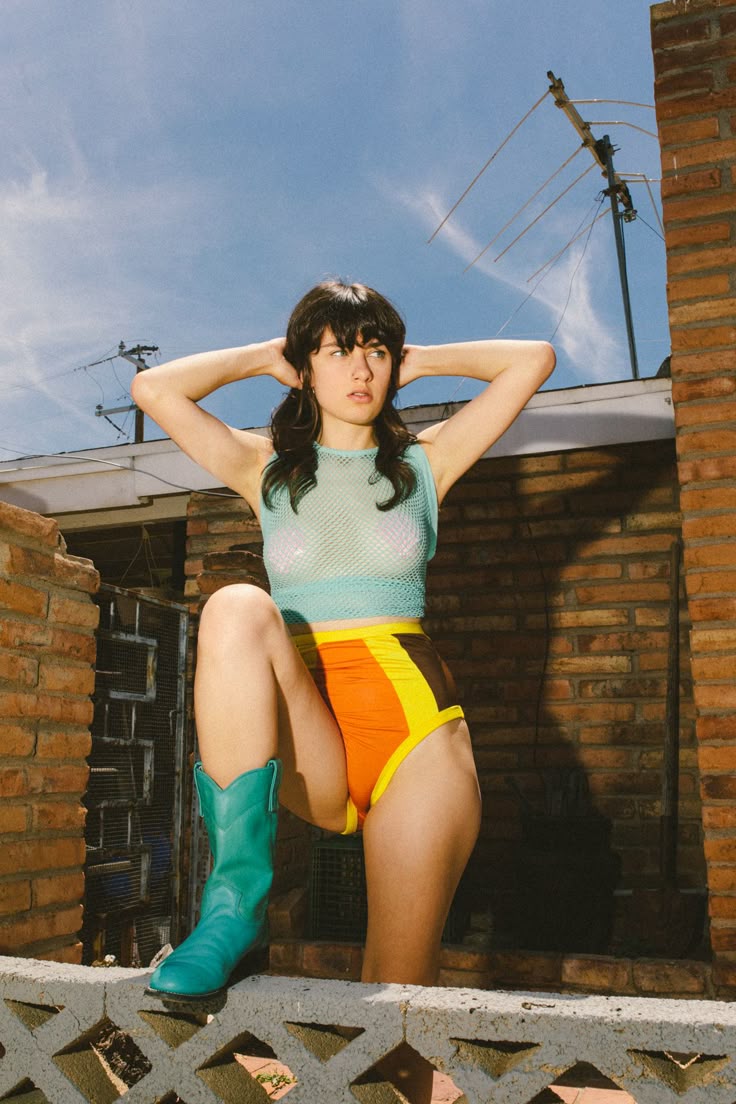 a woman is posing outdoors. she is wearing a light turquoise fishnet tank top, high waisted shorts with orange, yellow, and brown vertical stripes, and turquoise cowboy boots. she is standing on a concrete fence and leaning against a brick wall. in the background, there is an antenna against a blue sky.