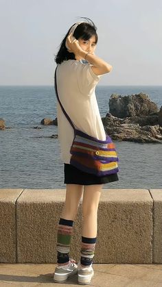 a woman stands on a seawall with her back to the camera. she is wearing a white tshirt, black skirt, colorful socks and white sneakers. she is wearing headphones and carrying a colorful striped bag. the sea is visible in the background with rocks emerging from the water. the photo is taken in natural daylight.  the woman is adjusting her headphones.