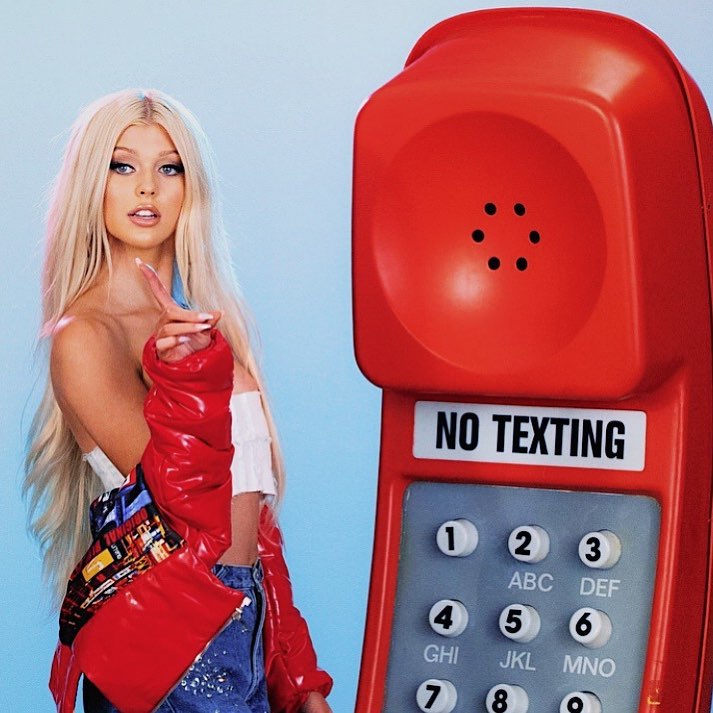 a young blonde woman in a red jacket and jeans is pointing with one hand towards the viewer, standing next to a large, red, button phone with a "no texting" label. the background is blue.