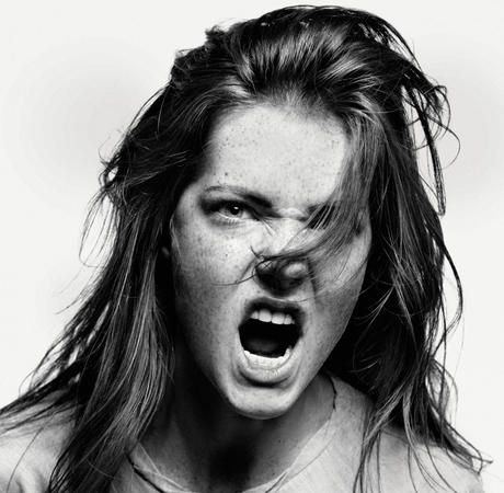 a black and white portrait of a young woman with long, messy hair. she is looking directly at the camera with a furious expression on her face. her mouth is wide open in a scream. the image is in focus and welllit.