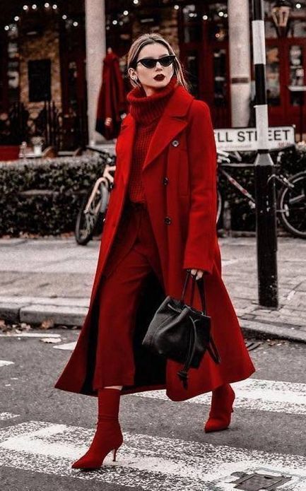a stylish woman in a headtotoe red outfit is crossing a city street.  she's wearing a long red coat, matching pants, and red ankle boots.  she carries a black bucket bag and is wearing sunglasses. the overall style is chic and elegant, with a monochrome color palette. the setting is a city street in autumn or winter.