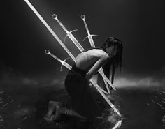 a woman sits with her back to the camera, kneeling amidst a cluster of swords. her body is illuminated by a single shaft of light, casting long shadows across the dark surface. the woman's expression is obscured by darkness, and the swords are arranged in a way that suggests violence, injury, and a possible struggle. the overall mood of the image is dark, dramatic, and mysterious.