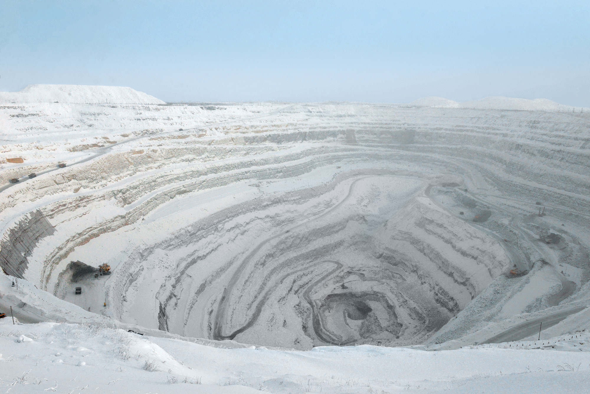 an aerial view of a large open pit diamond mine in siberia, russia. the mine is covered in snow and ice, with the walls of the pit showing the layers of rock that have been excavated.  a few vehicles can be seen at the bottom of the pit.  the sky is a clear blue, with some haze in the distance.