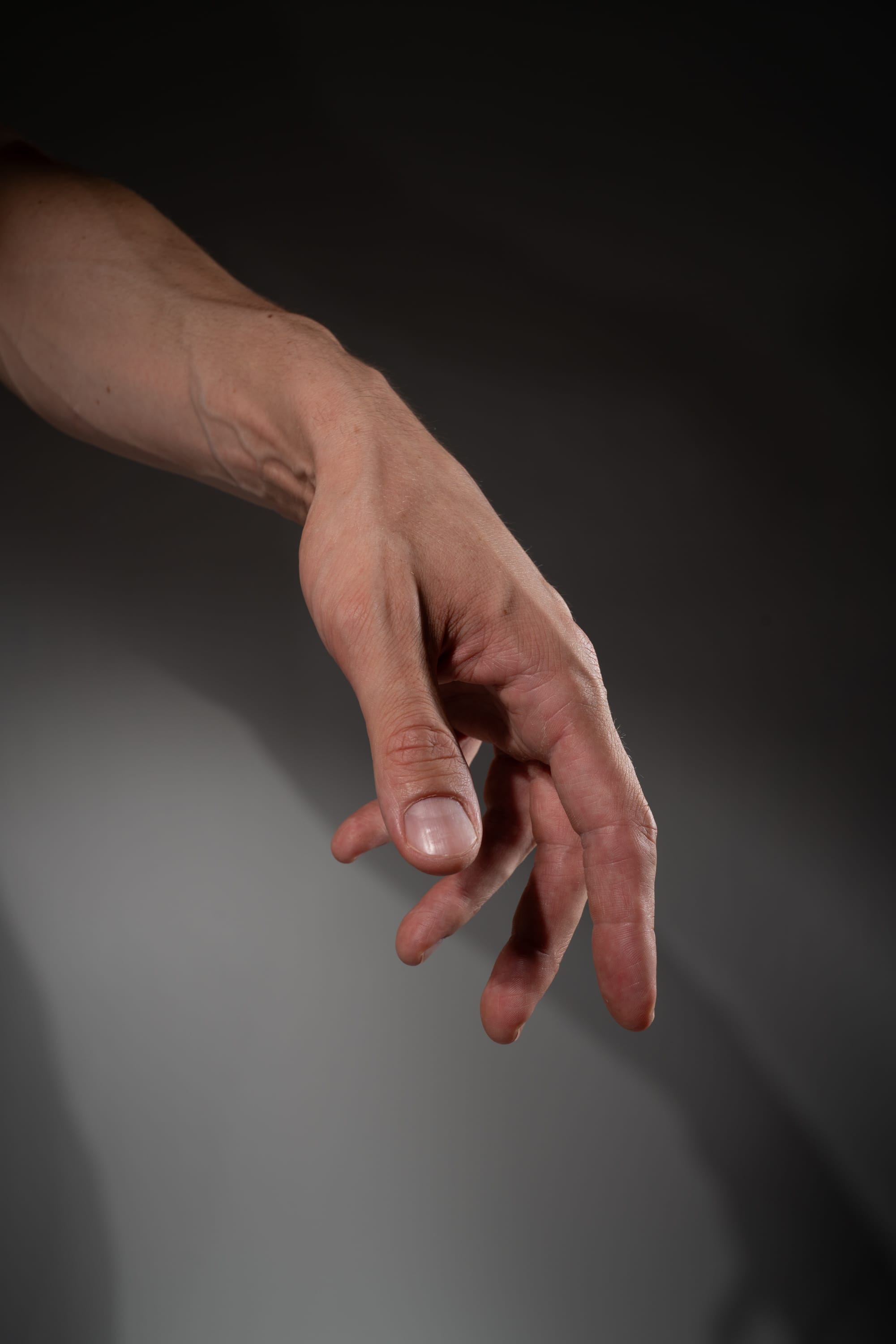 a man's right hand and forearm are extended downwards against a gray background, illuminated by soft studio lighting. the hand appears relaxed, with fingers slightly curled inward as if reaching towards something below the frame.