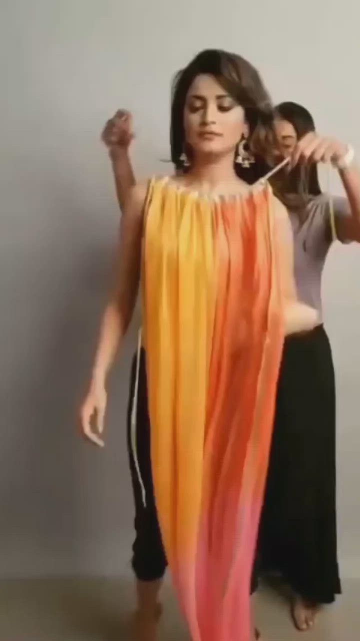 the image shows a woman wearing a long, flowing dress with a colorful gradient design. she has shoulderlength brown hair and is wearing earrings. two people, presumably stylists, are assisting her with the dress. the background is a plain grey color.