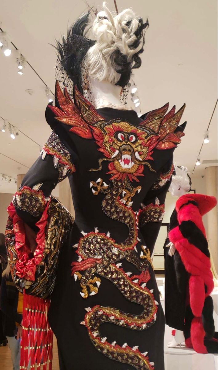 a black dress on a mannequin, featuring an elaborate dragon embroidery in red, gold, and white. the dress is displayed in an exhibition or museum setting, with other mannequins and exhibits visible in the background. the dragon design covers the back of the dress, from the shoulders down to the lower back, with the head positioned near the upper back.