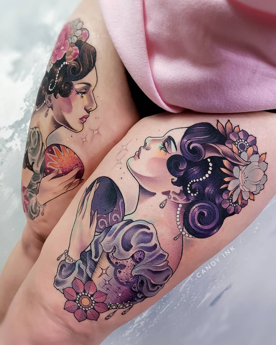 the image showcases two colorful neotraditional tattoos on someone's upper legs. each tattoo features a woman styled after art nouveau illustrations. the woman on the left, positioned on the outer thigh, holds a golden, stylized sun and wears flowers in her hair. the woman on the right, positioned on the front of the thigh, holds a dark, stylized moon and has purple hair with pearls and flowers woven in. both women wear elaborate clothing and jewelry. the artist's signature, "candy ink," is visible in black on the right thigh.