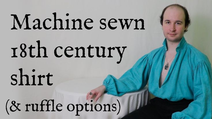 a man is wearing a machine sewn 18th century shirt with ruffle options. the shirt is blue. he is wearing a necklace and a ring. a table with a white tablecloth is to his left. the background is white. text on the left reads "machine sewn 18th century shirt (& ruffle options)".