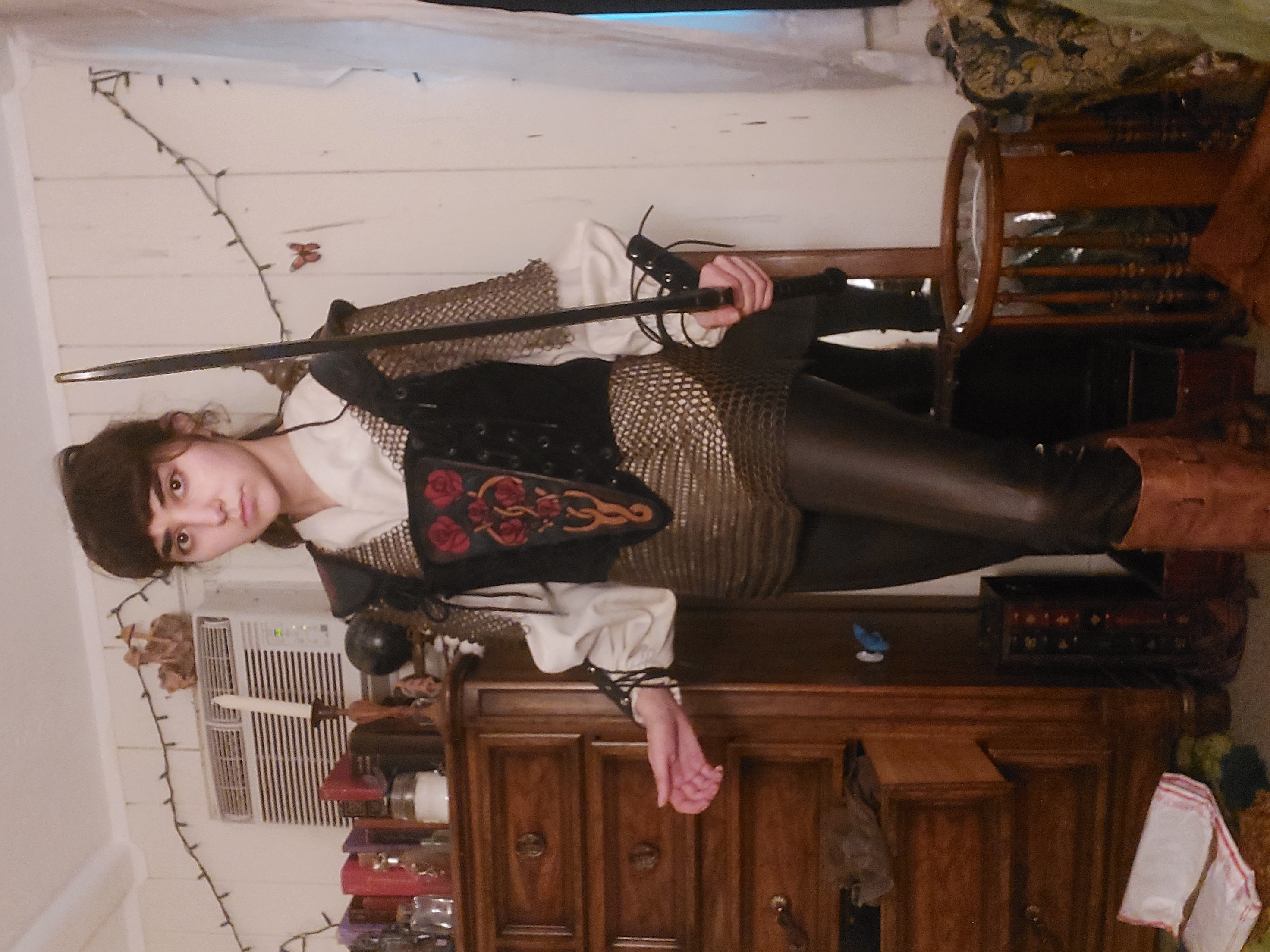 young woman dressed in a fantasy costume stands in a bedroom. her outfit includes a dark corset embroidered with roses and a celtic knot design, a chainmail shirt, a white tunic, dark leather pants, and brown boots. she holds a sword. the background includes wooden furniture and other decorative items.  the overall style is reminiscent of medieval or renaissance fantasy.