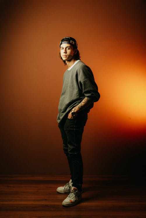 a full shot of a man standing indoors. he is wearing a hat, a sweatshirt, jeans, and sneakers. he has tattoos on his arm and is wearing a watch. he is looking at the camera. the background is a studio shot. the man is dressed in casual attire.