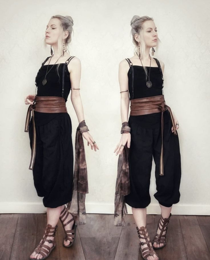 two images of a woman posing. she is wearing a black sleeveless top with thin straps, black harem pants, a wide brown wrap belt, and brown gladiator sandals. she has her blonde hair in an updo and is wearing jewelry including a necklace, earrings, and bangles. she is also holding a scarf. the background is a plain wall and a wooden floor. the images are in monochrome.