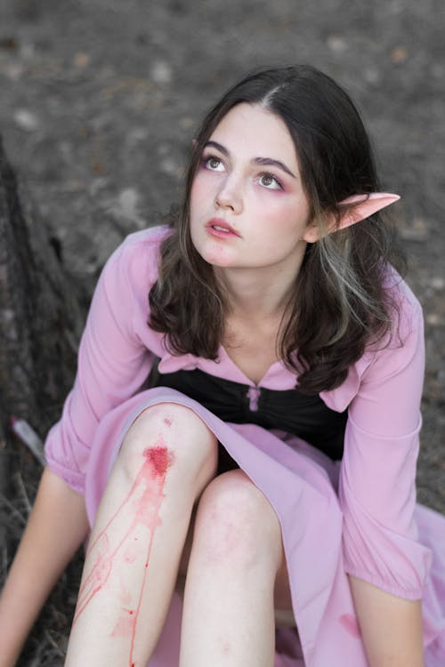 a woman dressed as an elf is sitting down. she is wearing a pink dress with a black bodice. she has elf ears and there is a wound on her knee with blood running down her leg. she is looking up.
