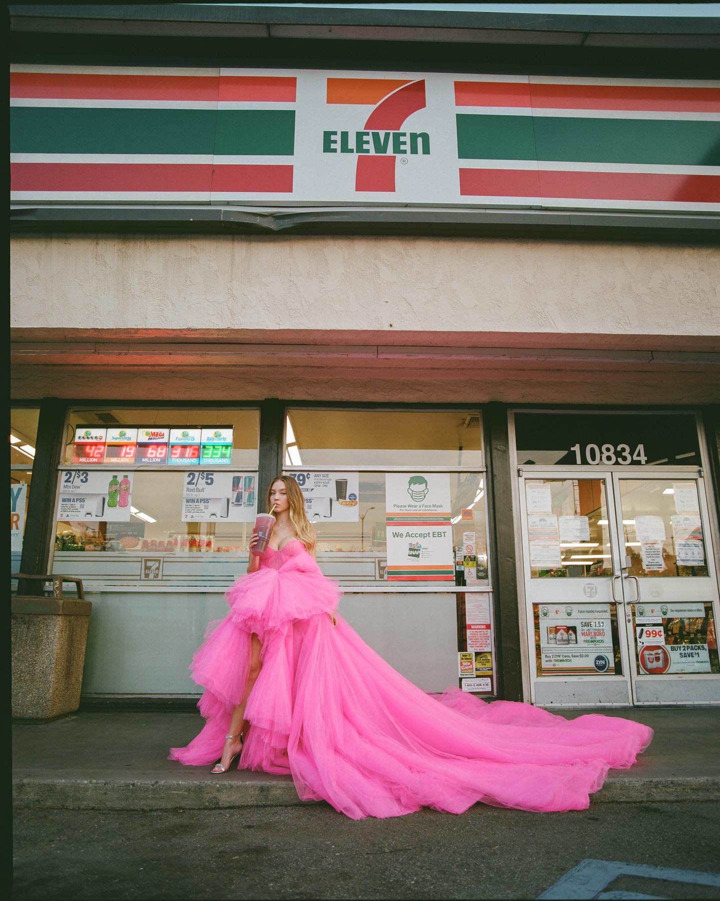 a young woman in a vibrant pink tulle highlow dress stands in front of a 7eleven convenience store.  the dress is voluminous and dramatic, creating a striking contrast against the everyday setting. she holds a drink in her hand. the store's signage and windows are clearly visible, and the number 10834 is displayed on the store's door. the overall aesthetic is a blend of high fashion and everyday life, suggesting a concept or editorial photoshoot.